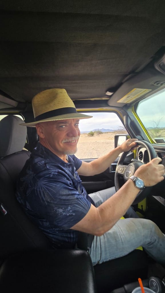 A candid photo of Rob Kaiser smiling behind the wheel of his High Velocity yellow Jeep; he’s wearing a blue floral shirt and a straw fedora, with the desert landscape visible through the window.