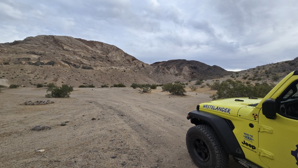 A partial view of the yellow Jeep's hood and front fender in the foreground, looking out toward a faint trail winding through scrub brush toward rugged, layered desert hills under an overcast sky.