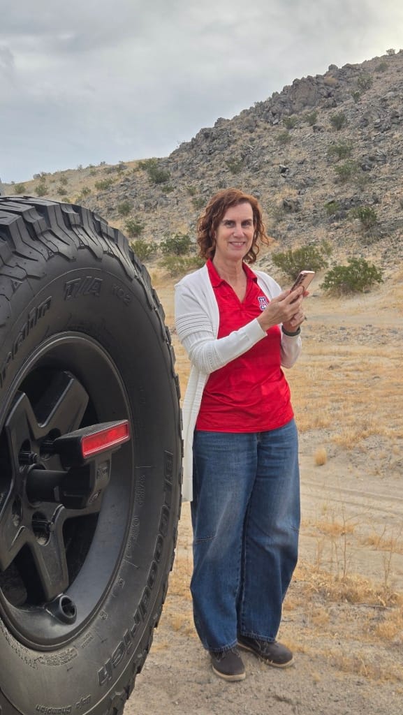 Lynn Kaiser stands next to a large 35-inch Jeep tire, wearing a red University of Arizona polo and a white cardigan, checking her phone against a backdrop of rocky desert hills.