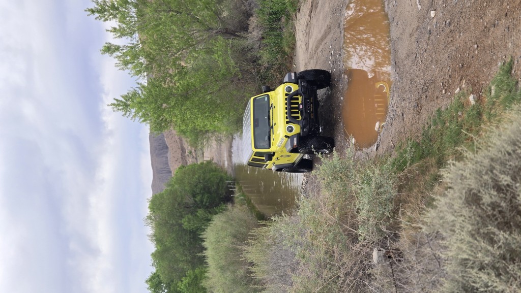 High Velocity yellow Jeep Wrangler navigating a muddy section of the trail at a large water-filled riverbed crossing, surrounded by green trees and desert brush.