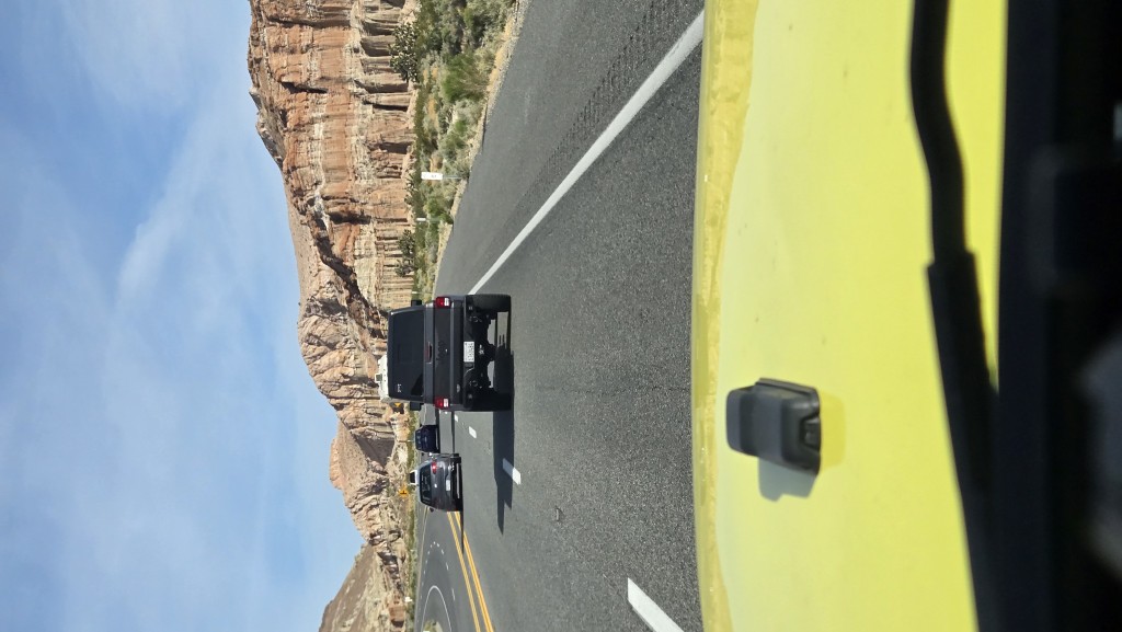 A paved highway through a desert landscape dominated by massive, red rock cliffs under a pale blue sky.