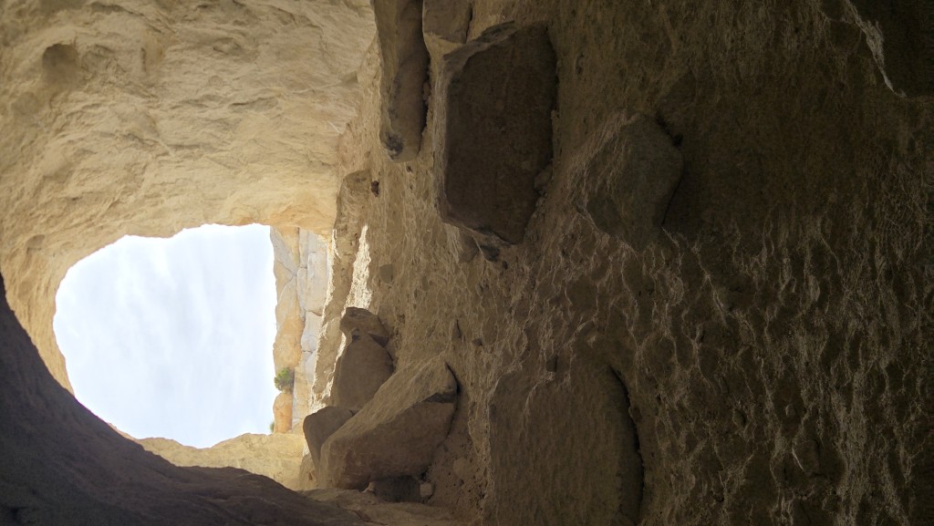 A view looking out from inside a dark mine entrance toward a bright desert landscape. The jagged, dark silhouette of the cave opening frames the sunlit dirt trail and distant hills outside.
