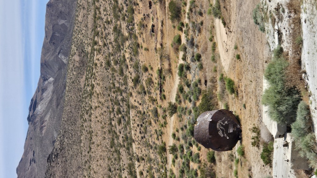A close-up of a weathered, rusted metal bin or machinery part resting on rocky desert ground with modern graffiti art.