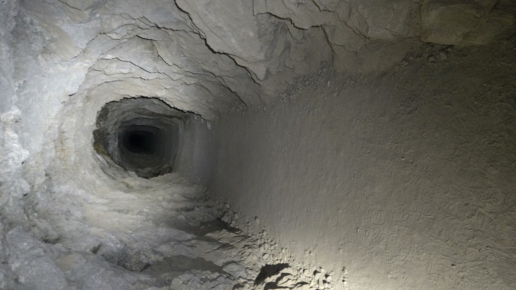 A long, narrow tunnel carved through rough, light-colored rock. The path is dusty and flat, disappearing into total darkness in the distance, illuminated only by a nearby light source.