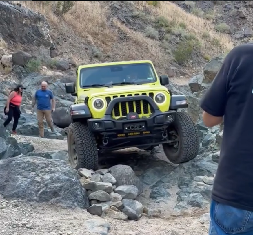 A High Velocity yellow Jeep Wrangler navigating a narrow, rocky canyon trail. One front tire is significantly articulated over a large boulder while a spotter and bystanders watch the progress closely.